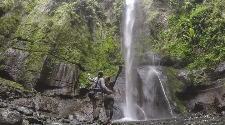 A small group standing at the base of a tall waterfall in a mossy rock gorge