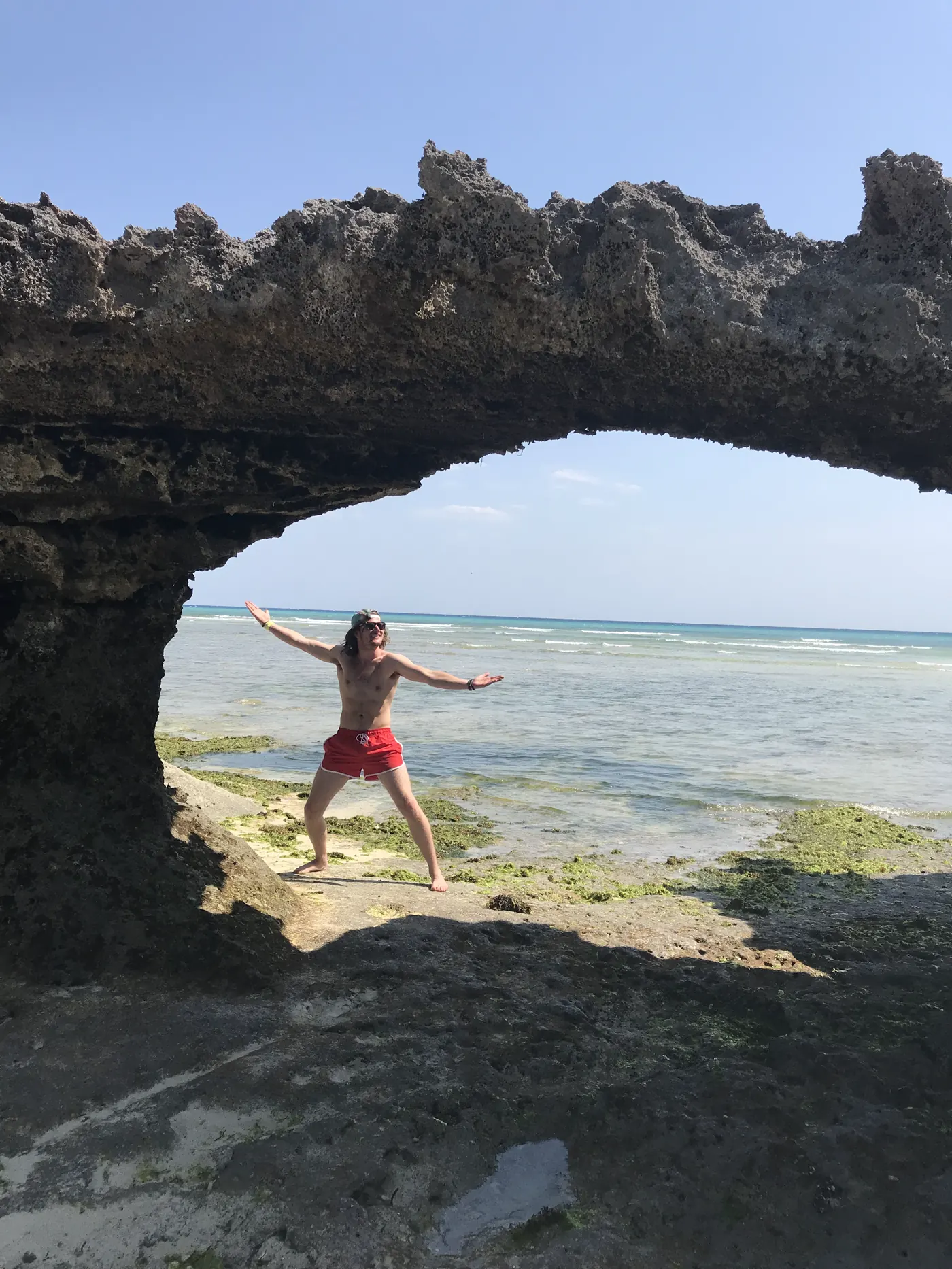 A person posing under a natural limestone rock arch above turquoise water