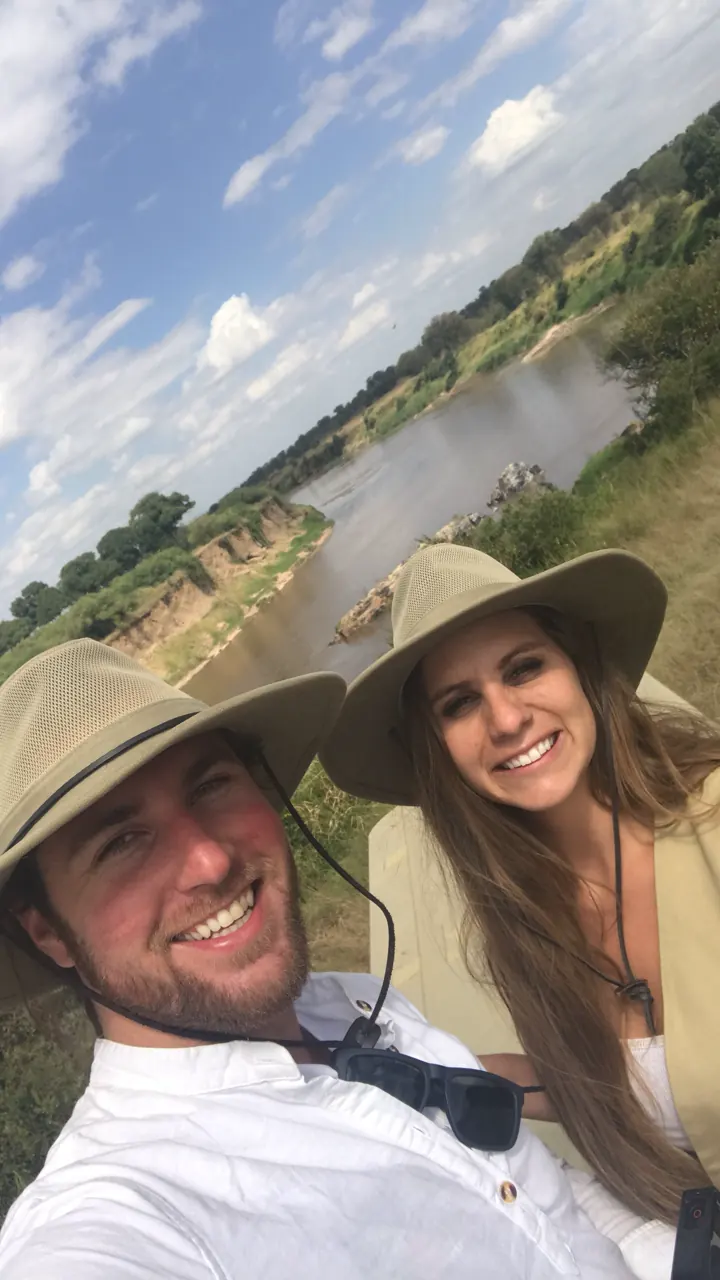 A smiling couple in safari hats taking a selfie above a river valley