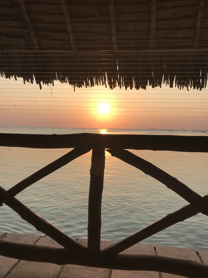 The sun setting over the ocean seen from a thatched balcony with wooden railings