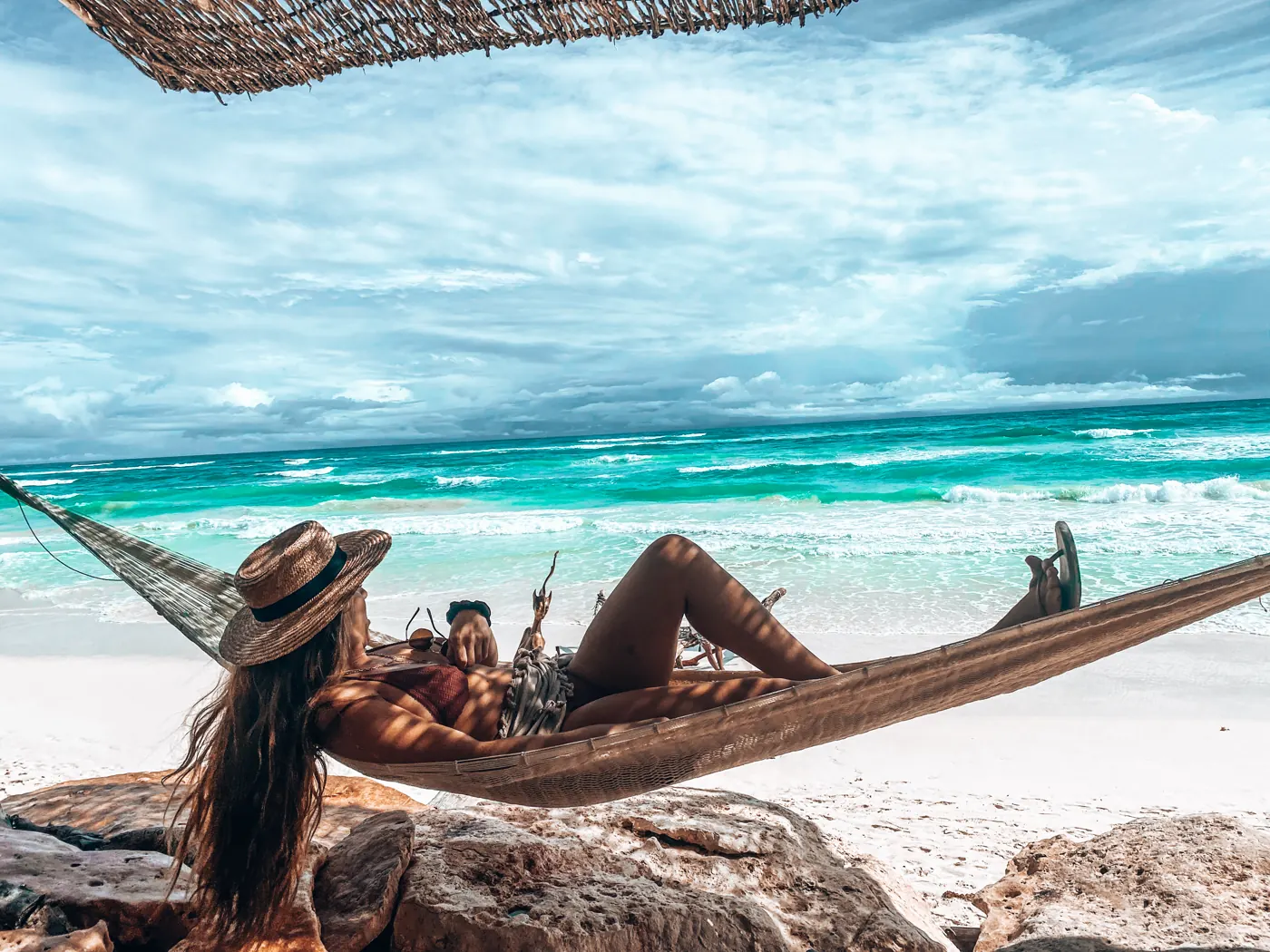 A woman in a sun hat lounging in a hammock above white sand and turquoise sea
