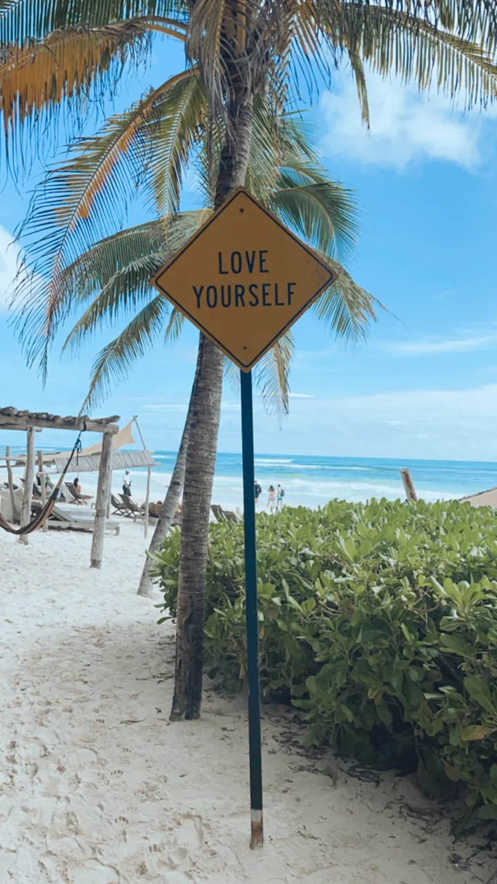 A yellow road sign reading Love Yourself on a sandy beach path under palms