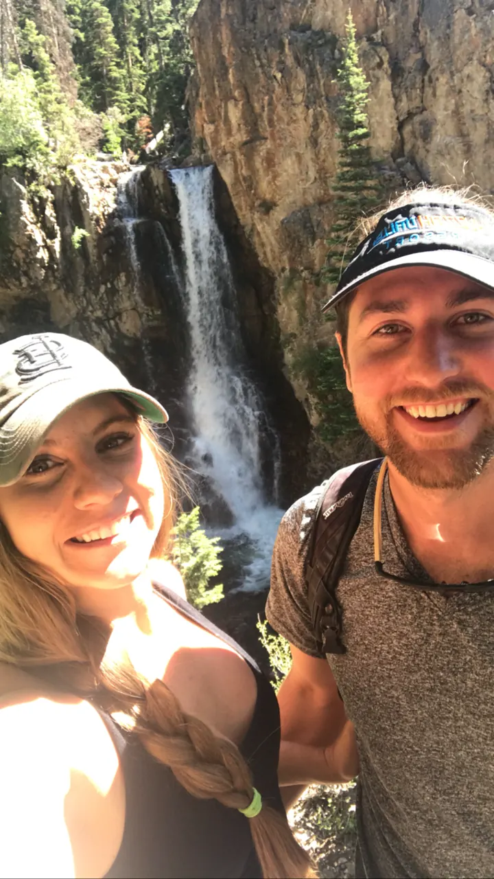 A couple in hats taking a selfie in front of a waterfall in a red-rock canyon