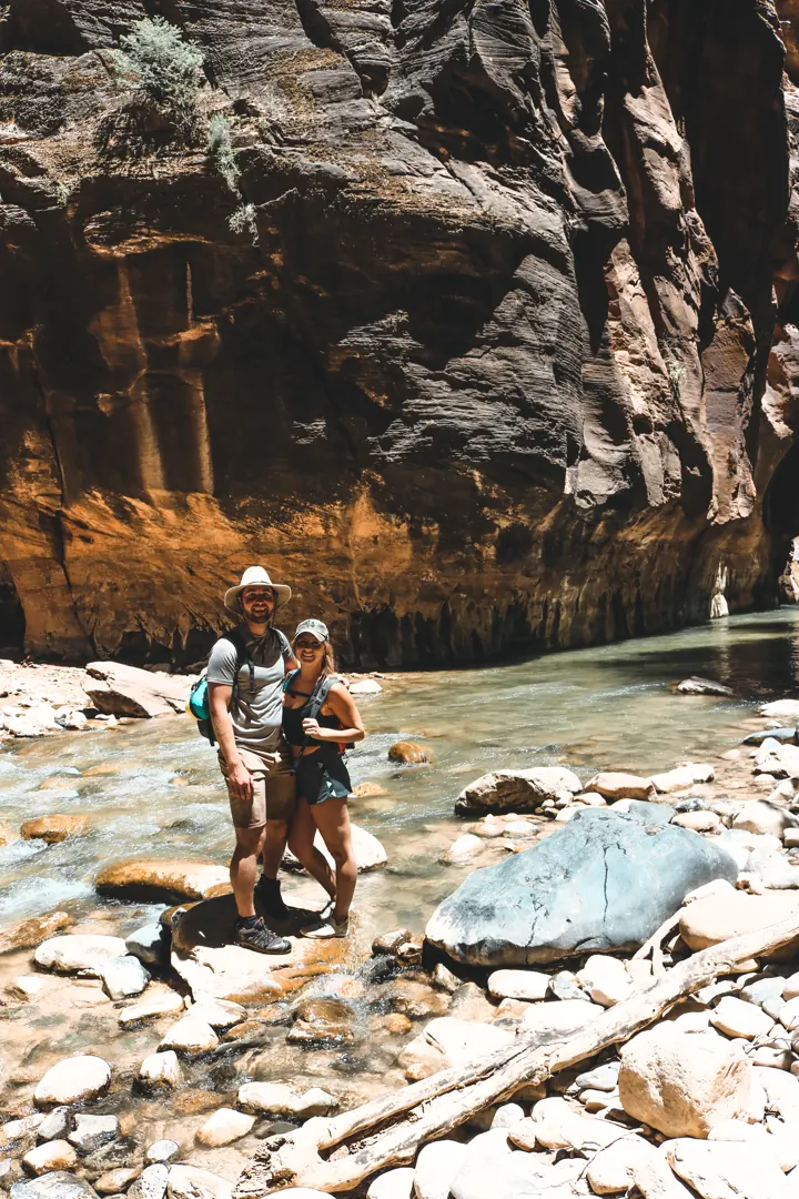 A couple standing on rocks by the river in The Narrows between towering canyon walls